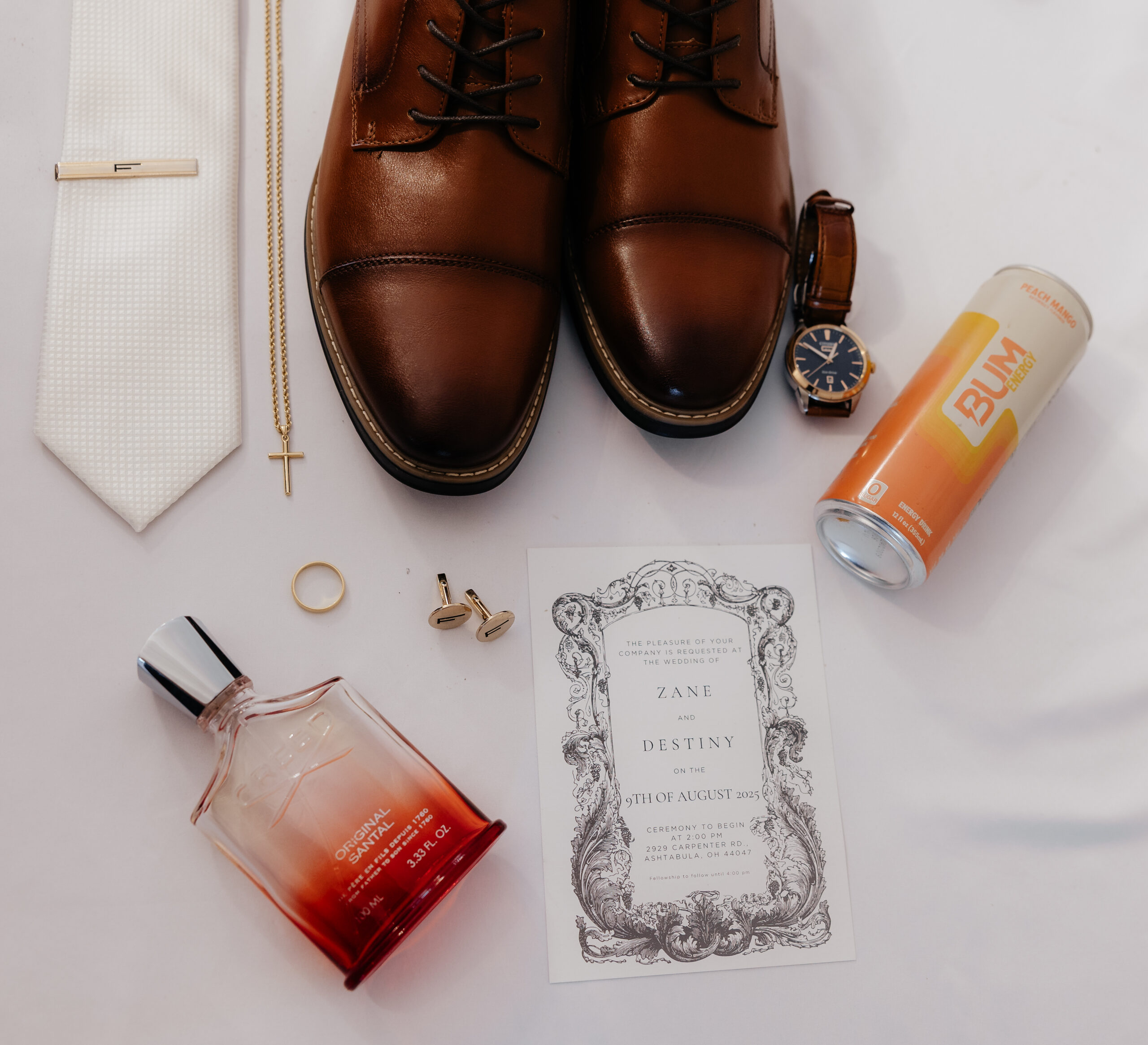 A flat lay detail photo of a groom's items including shoes, watch, cologne, and their wedding invitation.