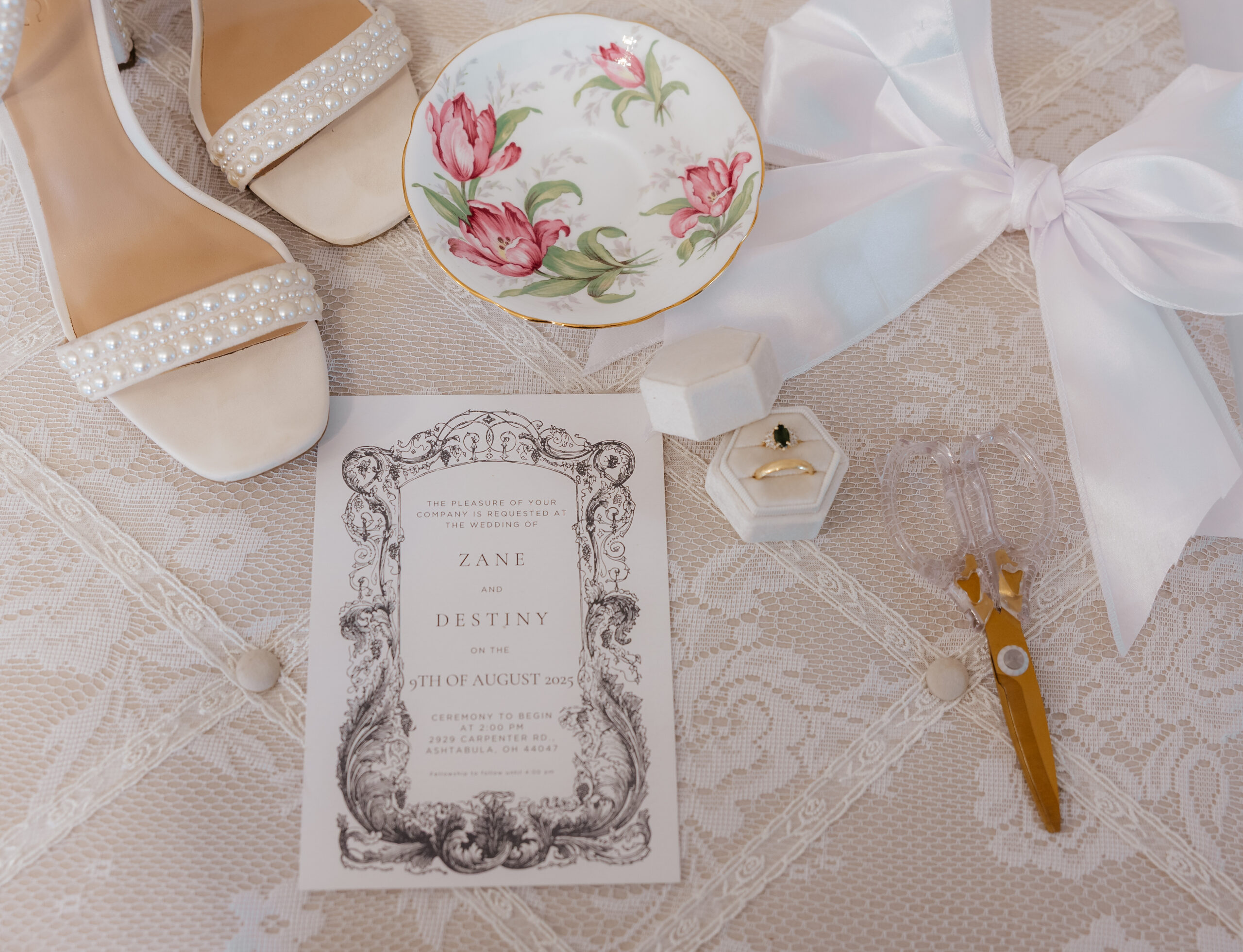 A flat lay detail photo of a bride's items including shoes, a bow, and a ring dish, and their wedding invitation.