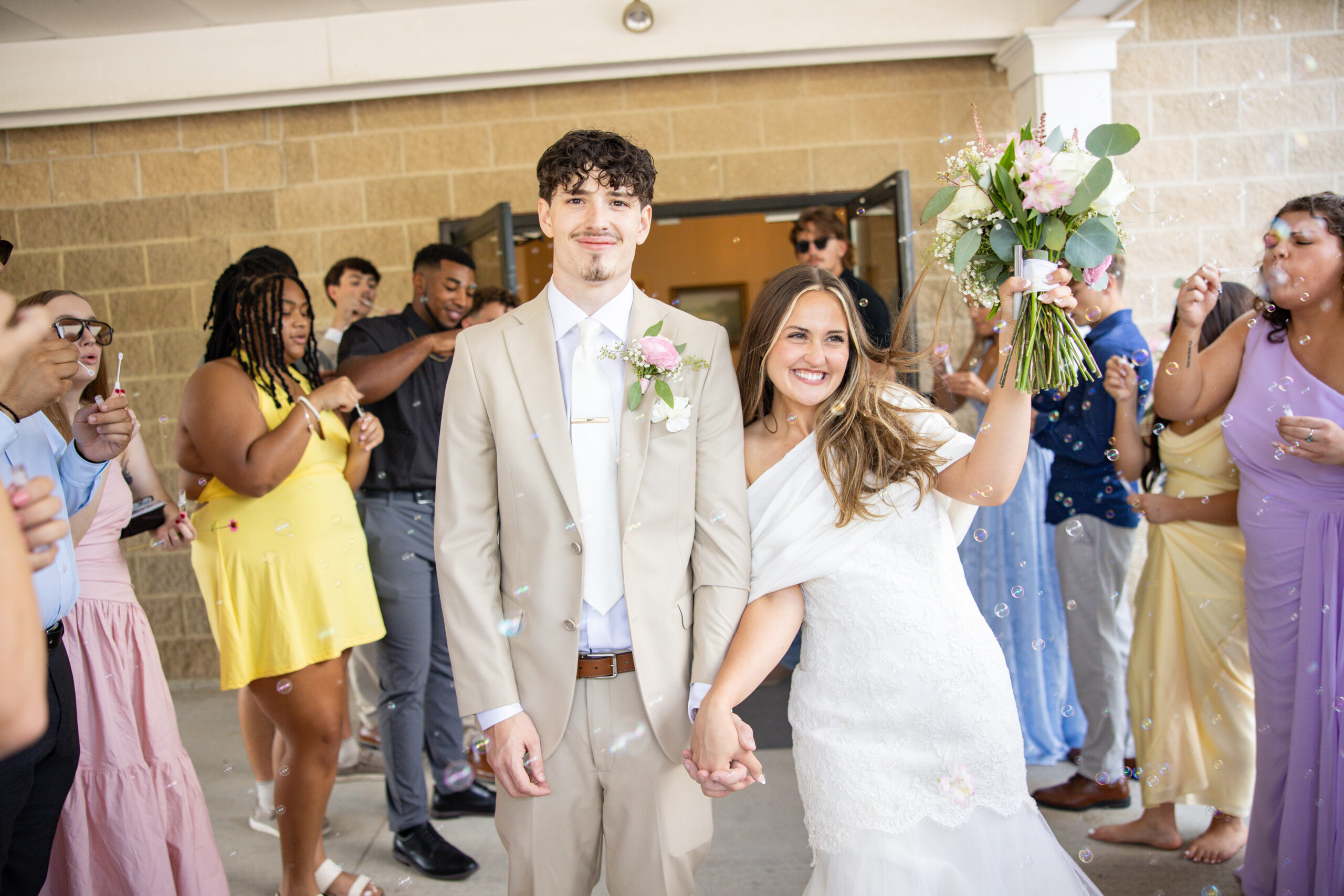 A bride and groom walking down an aisle of family and friends as bubbles are being blown at them.